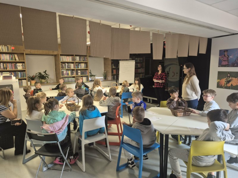 Grupa dzieci siedzi przy stolikach i patrzy w stronę stojącej na środku bibliotekarki, która pokazuje im otwartą encyklopedię