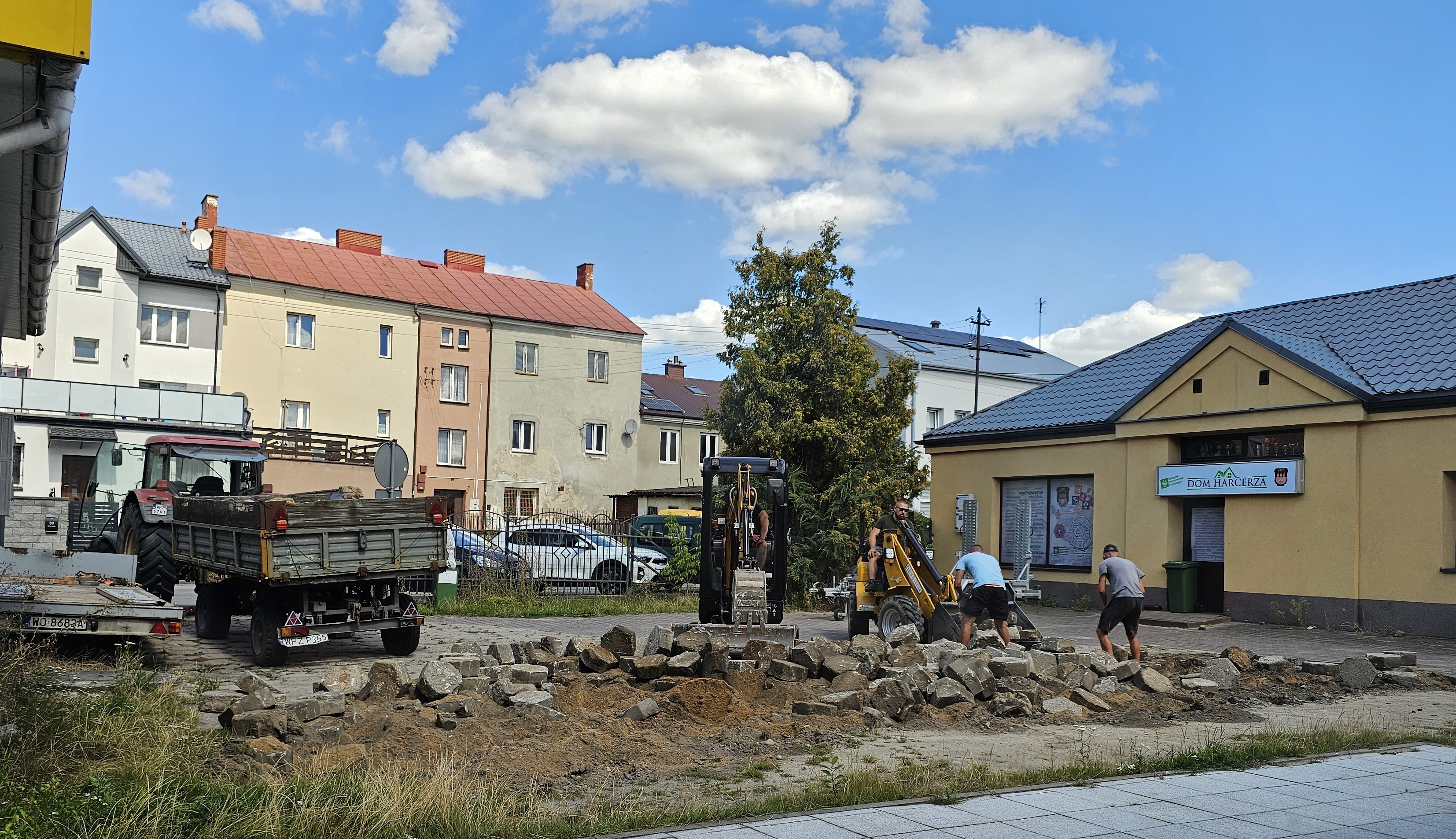 Prace remontowe na placu przed budynkiem Domu Harcerza. Widoczni są robotnicy pracujący przy zerwanej nawierzchni oraz ciężki sprzęt budowlany: minikoparka, ładowarka i traktor z przyczepą