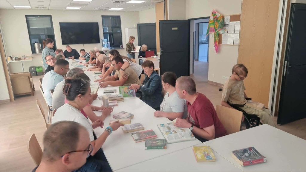 Szerokie ujęcie jaśniejszej sali bibliotecznej lub warsztatowej. Przy dwóch połączonych, białych stołach siedzi kilkunastu uczestników spotkania. Przed nimi rozłożone są liczne książki. Część osób rozmawia ze sobą, inne przeglądają strony. Po prawej stronie przy stole siedzi osoba na wózku inwalidzkim