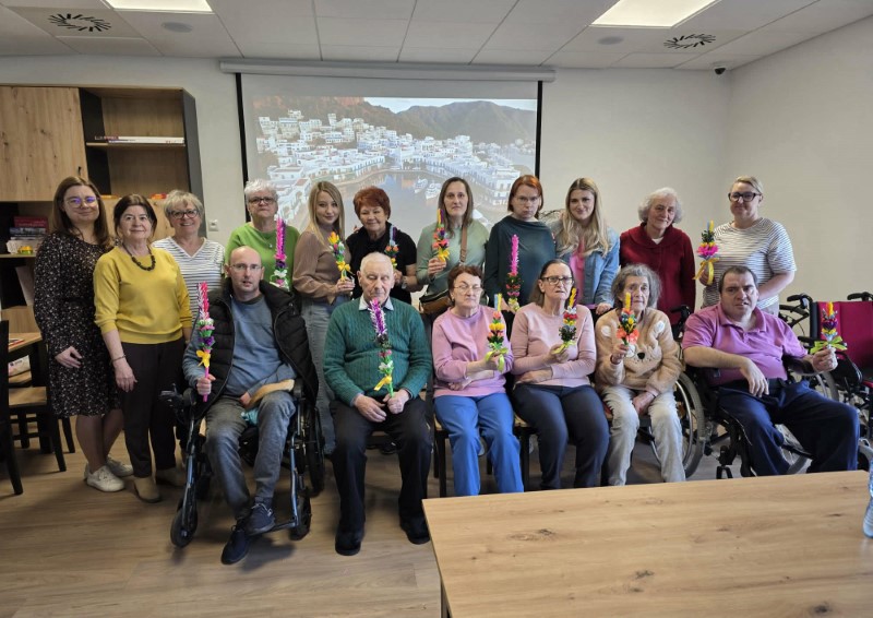 Na zdjęciu widać grupę dorosłych uśmiechniętych osób pozujących do zdjęcia. Każda osoba trzyma w dłoni własnoręcznie stworzoną palmę wielkanocną