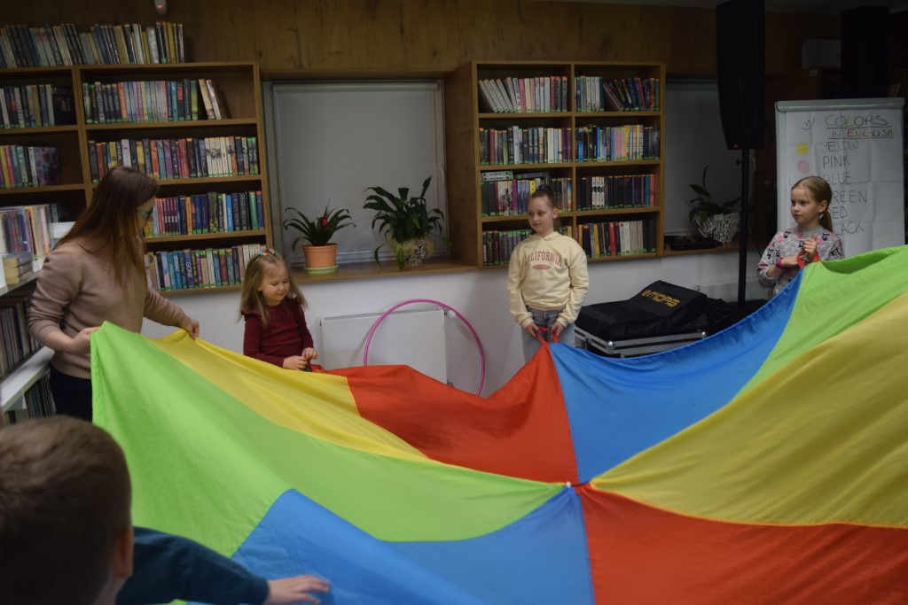 Dzieci wraz z bibliotekarką bawią się w bibliotece dużą, kolorową chustą animacyjną. Chusta jest rozłożona i trzymana przez uczestników na różnych wysokościach. W tle znajdują się regały wypełnione książkami oraz rośliny doniczkowe na parapecie