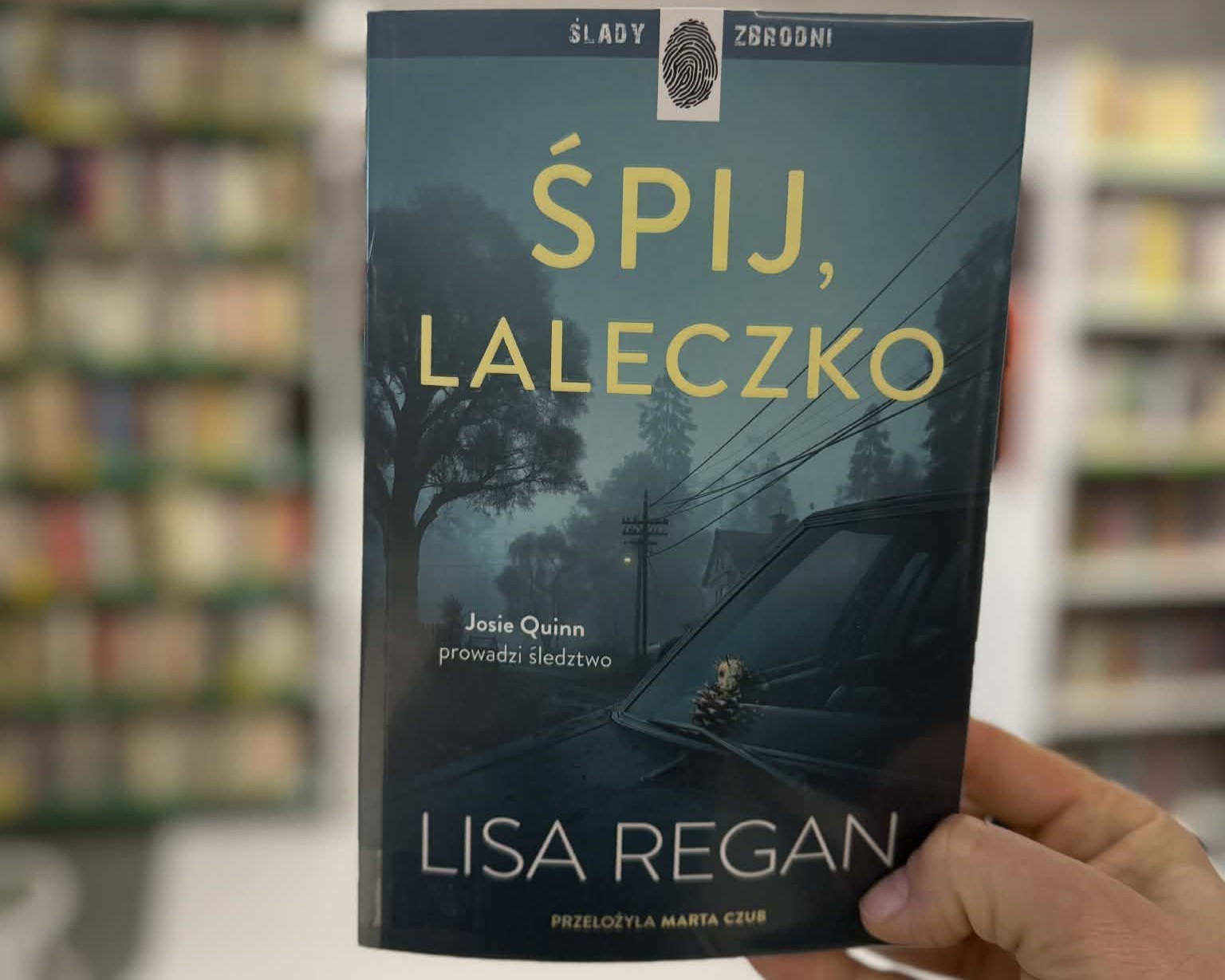 Na pierwszym planie widnieje męska dłoń trzymająca książkę Lisy Regan "Śpij, laleczko". W tle widać rozmyte regały z książkami. Zdjęcie zrobione jest w bibliotece