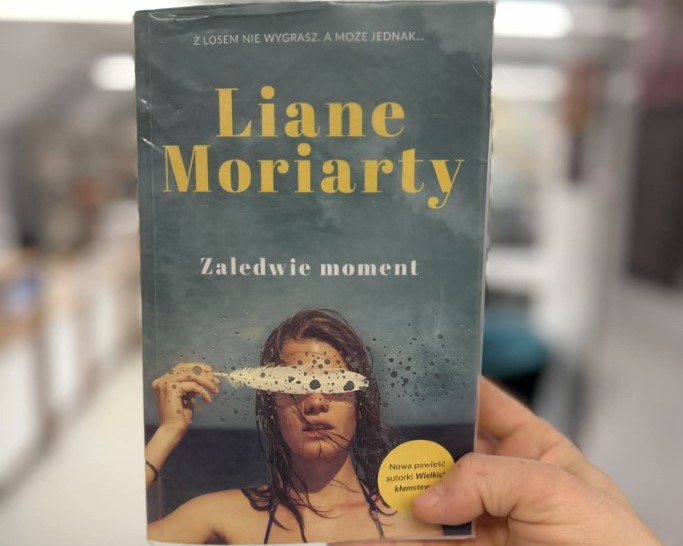 Na pierwszym planie widnieje męska dłoń trzymająca książkę Liane Moriarty "Zaledwie moment". W tle widać rozmyte regały z książkami. Zdjęcie zrobione jest w bibliotece