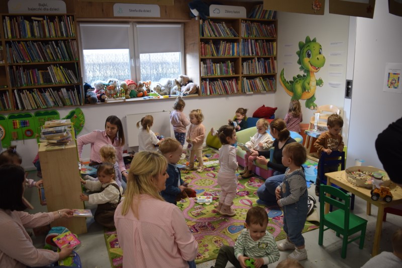 W centralnej części zdjęcia widać dużą grupę trzylatków, które bawią się na kolorowym dywanie w kąciku dla dzieci. Towarzyszą im opiekunki, które siedzą z nimi na wydanie i pokazują im książeczki 