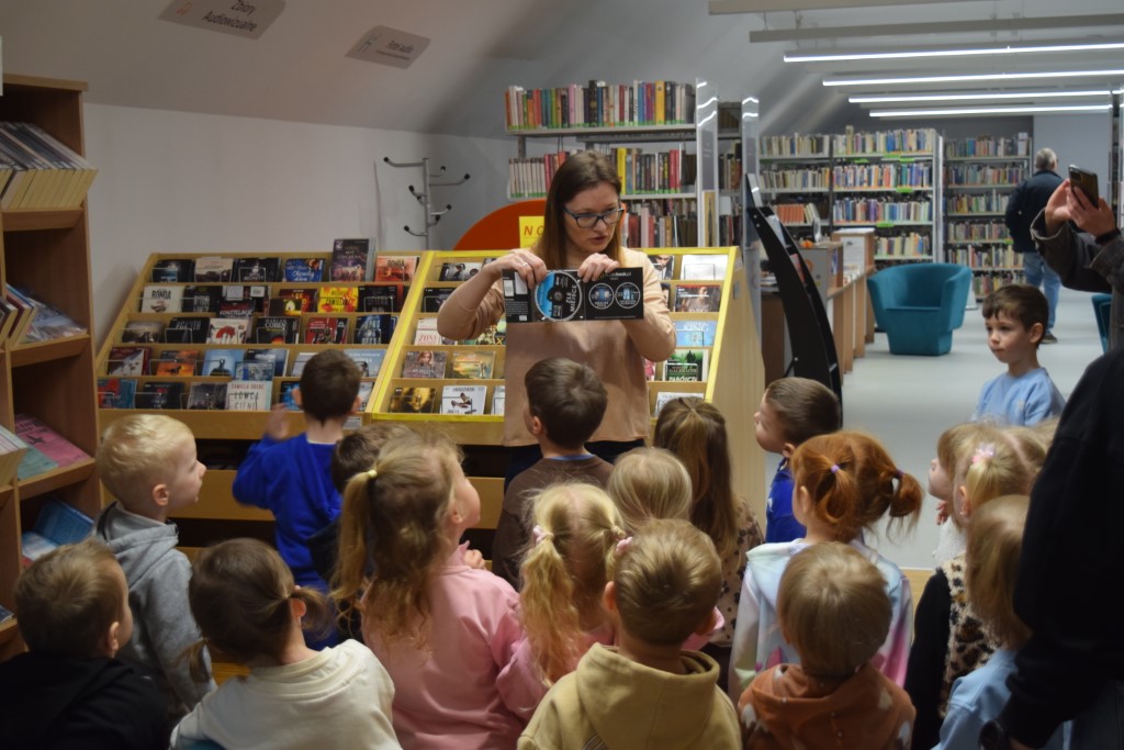 Bibliotekarka w okularach stoi przed grupą małych dzieci w dziale zbiorów audiowizualnych. Kobieta pokazuje dzieciom otwarte opakowanie z płytą CD, tłumacząc zasady wypożyczania multimediów. Dzieci z zaciekawieniem patrzą na prezentację