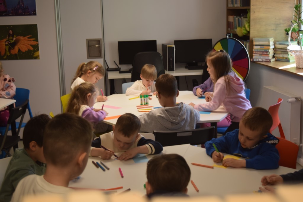 Grupa dzieci siedzi przy dużym, białym, okrągłym stole w jasnym pomieszczeniu. Dzieci rysują kredkami na kartkach papieru. W tle widoczne są stanowiska komputerowe oraz półki z książkami