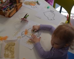 Przy stoliku siedzi trzylatka. Przed nią leży kartka z ilustracją kurczaka, na którym dziewczynka rozsypała ziarna. W tle widać materiały plastyczne oraz prace innych dzieci