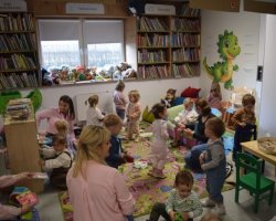 W centralnej części zdjęcia widać dużą grupę trzylatków, które bawią się na kolorowym dywanie w kąciku dla dzieci. Towarzyszą im opiekunki, które siedzą z nimi na wydanie i pokazują im książeczki