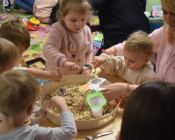 Grupa dzieci siedzi wokół stolika, na którym leży taca z nasionami. Dzieci trzymają w dłoniach łyżki, za pomocą których nakładają ziarna i nasiona w plastikowe kubki, które imitują wielkanocne zwierzęta