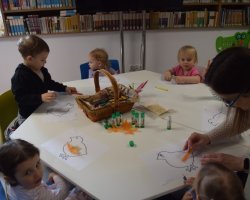 Wokół stolika, na którym są materiały plastyczne siedzą trzylatki. Przed każdym dzieckiem znajduje się kartka z wydrukowaną kolorowanką kurczaka. Bibliotekarka pomaga jednej z dziewczynek przykleić na kartkę ozdobne piórko