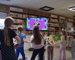Grupa dzieci stoi odwrócona plecami do fotografa. Naśladują ruchy taneczne wyświetlane na dużym ekranie telewizora, gdzie widoczna jest kolorowa, pop-artowa grafika przedstawiająca usta. W tle znajdują się wysokie, drewniane regały wypełnione książkami