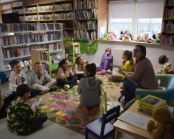 W kąciku dla dzieci, klasa siedzi w kole na dywanie i słucha bibliotekarki prowadzącej zajęcia o tematyce wielkanocnej. 