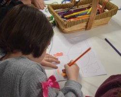 Dziewczynka koloruje szablon kury. Na stoliku znajdują się kleje oraz koszyk z kolorowymi kredkami.