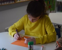 Dziewczynka koloruje szablon kury. Przed nią na stoliku stoją kleje w sztyfcie oraz leżą nożyczki.