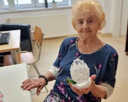 W centralnej części zdjęcia znajduje się uśmiechnięta kobieta, która trzyma prezentuje w dłoni gotową ozdobę wielkanocną. Za nią po lewej stronie stoi komputer dla użytkowników. Po prawej stronie zdjęcia znajduje się brązowa witryna.