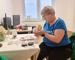 Kobieta w niebieskiej bluzce siedzi przy stole, gdzie tworzy wielkanocne ozdoby z akrylowych jajek. Na stole przed nią znajdują się materiały potrzebne do stworzenia pracy.