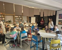 Zdjęcie przedstawia grupę dzieci, które siedzą przy białych stolikach. wszystkie są zwrócone do bibliotekarek, które prowadzą zajęcia
