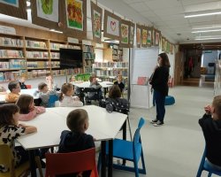 Grupa dzieci siedzi przy stolikach i patrzy w stronę stojącej na środku bibliotekarki, która pokazuje im otwartą encyklopedię