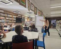 Bibliotekarka wskazuje na tablicę, gdzie spisane są warunki bezpiecznego korzystania z Internetu. W centralnej części zdjęcia widać dzieci przy stolikach