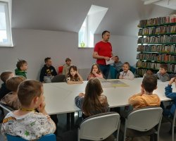 Bibliotekarz opowiada dzieciom ciekawostki o książkach. Klasa siedzi przy dużym stole, na którym leżą książki różnych formatów