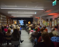 Perspektywiczny widok na długą salę Miejskiej Biblioteki Publicznej w Przasnyszu wypełnioną publicznością. Po lewej stronie znajdują się regały z książkami, po prawej galeria rysunków, a na końcu sali, na podwyższeniu, trwa spotkanie z Arturem Dziurmanem