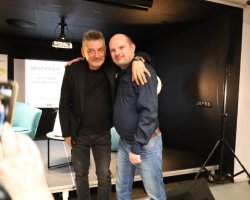 Artur Dziurman pozuje na scenie do zdjęcia z uśmiechniętym mężczyzną w niebieskiej koszuli w kropki i dżinsach, obejmując go ramieniem. Na pierwszym planie z lewej strony widoczna jest dłoń innej osoby robiącej im zdjęcie smartfonem