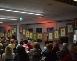 Publiczność siedząca na krzesłach w sali bibliotecznej, sfotografowana od tyłu podczas spotkania. Wzdłuż prawej ściany, na sznurku z klamerkami, rozwieszona jest wystawa kolorowych prac plastycznych wykonanych przez uczestników Środowiskowego Domu Samopomocy oraz członkiń Klubu Rekodzieła