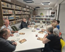 Widok z boku na uczestników dyskusji siedzących przy białym stole konferencyjnym w bibliotece. Przed kilkoma osobami leżą egzemplarze tej samej książki z widoczną ilustracją na okładce. Wszyscy są zwróceni ku sobie, zaangażowani w rozmowę. Tło stanowią regały z książkami oraz fragment strefy dziecięcej z zielonym smokiem na ścianie