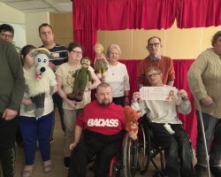 Grupa osób pozuje do zdjęcia na tle czerwonej kurtyny teatrzyku pacynkowego. Uczestnicy trzymają wykonane ręcznie pacynki przedstawiające wilka, żołnierza, lalkę i wiewiórkę. Dwóch mężczyzn na pierwszym planie porusza się na wózkach inwalidzkich; jeden z nich trzyma pamiątkowy bilet z napisem „Teatrzyk pacynkowy – Czerwony Kapturek”