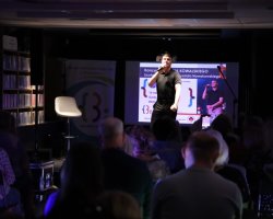 Damian Kowalski podczas koncertu w przyciemnionej sali bibliotecznej. Wokalista dynamicznie śpiewa na scenie, trzymając mikrofon. W tle widoczny jest ekran z plakatem koncertu, a przed sceną siedzą widzowie, z których jeden nagrywa występ telefonem