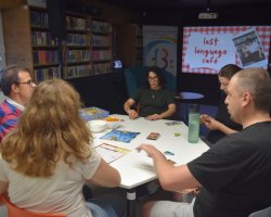 Uczestnicy językowego spotkania przy planszówkach siedzą przy białym stole w sali bibliotecznej. Perspektywa ujęcia ukazuje z bliska dwoje uczestników z lewej strony oraz mężczyznę po prawej, który sięga po kartę do gry. W tle widoczne są regały z książkami oraz ekran projektora z grafiką "last language café"
