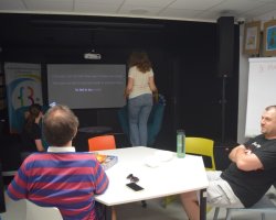 Uczestnicy spotkania językowego w bibliotece siedzą wokół białego stołu w multimedialnej sali o ciemnych ścianach. Jedna z kobiet stoi tyłem do obiektywu przed dużym ekranem projektora, na którym wyświetlany jest tekst piosenki w języku angielskim. Po prawej stronie widoczny jest mężczyzna siedzący z założonymi rękami, a obok niego stoi biała tablica z napisem "HANGMAN"