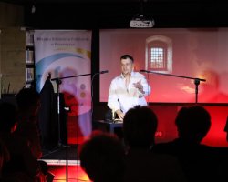 Aktor w białej koszuli występuje na scenie przed mikrofonem, gestykulując dłonią. W tle widoczny jest ekran projekcyjny z ilustracją przedstawiającą okno z kratą oraz roll-up Miejskiej Biblioteki Publicznej w Przasnyszu. Na pierwszym planie widoczne są sylwetki zgromadzonej publiczności
