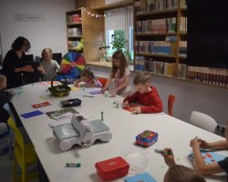 Grupa dzieci siedzi przy dużym białym stole w bibliotece i wykonuje kartki okolicznościowe metodą scrapbookingu. Na stole leżą kolorowe papiery, kleje w sztyfcie i maszynka do wycinania wzorów. W tle widać regały z książkami oraz uśmiechniętą instruktorkę rozmawiającą z dziećmi