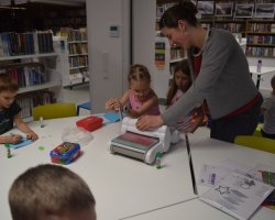 Zbliżenie na wolontariuszkę pomagającą dziewczynce w obsłudze białej maszynki do scrapbookingu. Dziewczynka kręci korbką, aby wyciąć kształty w papierze. Obok na stole inne dziecko przykleja elementy do swojej kartki. W tle znajdują się biblioteczne regały wypełnione książkami