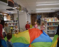 Grupa dzieci i bibliotekarka trzymają za brzegi dużą, kolorową chustę animacyjną w przestrzeni bibliotecznej. Wszyscy są uśmiechnięci i przygotowani do wspólnej zabawy ruchowej