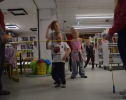 Dzieci biorą udział w zabawie pod nazwą "limbo" w bibliotece. Bibliotekarka i wolontariuszka trzymają rozciągnięty żółty sznur, pod którym przechodzi uśmiechnięty chłopiec w białej koszulce. Pozostałe dzieci czekają w kolejce, obserwując zabawę
