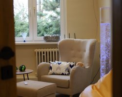 Widok przez otwarte drzwi na pokój wyciszeń w Filii nr 1. W centrum znajduje się beżowy fotel z poduszką i królikiem obciążeniowym, obok kolumna świetlna i okno z widokiem na drzewa iglaste. Na parapecie stoi radioodtwarzacz