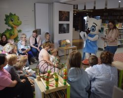 Grupa małych dzieci wraz z opiekunami siedzi w kręgu w kąciku dziecięcym w bibliotece. Wszyscy z uwagą patrzą na postać w dużym kostiumie maskotki Kici Koci, która stoi przed nimi w niebieskiej sukience i białych rękawiczkach. Obok maskotki stoi kobieta trzymająca otwartą książkę. Na ścianie po lewej stronie widnieje duża naklejka z zielonym smokiem, a na podłodze leży kolorowa mata z zabawkami