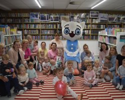 Pamiątkowe zdjęcie grupowe uczestników spotkania w bibliotece. Ponad piętnaścioro małych dzieci oraz kilka dorosłych kobiet siedzi i klęczy na pasiastym dywanie. W samym centrum grupy stoi maskotka Kici Koci w niebieskiej sukience. W tle znajdują się wysokie regały wypełnione książkami, nad którymi zawieszono galerię zdjęć. Na pierwszym planie dziewczynka trzyma różowy balon