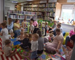 Swobodna zabawa dzieci i opiekunów po spotkaniu w bibliotece. Maluchy siedzą na kolorowym dywanie, układają plastikowe klocki z dużego pojemnika i bawią się zabawkami edukacyjnymi. Dorośli siedzą obok nich na podłodze lub na niskich, kolorowych pufach. W tle widoczne są regały z literaturą dziecięcą oraz duże okno, na którym stoją pluszowe maskotki
