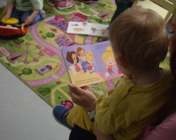 Widok zza ramienia małego dziecka siedzącego na kolanach dorosłego i oglądającego otwartą, kolorową książeczkę obrazkową przedstawiającą ilustrację z radosną rodziną