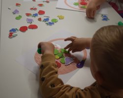 Zbliżenie na rączki dziecka przy białym stoliku, które przykleja kolorowe, piankowe naklejki w kształcie owoców – truskawek, winogron i jabłek – na kartkę z rysunkiem koszyka