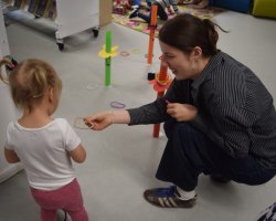Wolontariuszka w koszuli w paski kuca na dywanie i podaje kolorową obręcz małej dziewczynce spiętej w dwa kucyki. Wokół na podłodze rozstawione są plastikowe stojaki do gry zręcznościowej