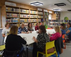 Grupa ośmiu kobiet siedzi przy dużym, białym stole w nowoczesnej sali bibliotecznej. Kobiety rozmawiają, piją kawę i herbatę. W tle widoczne są wysokie, drewniane regały wypełnione książkami oraz kolorowe krzesła. Jedna z uczestniczek porusza się na wózku 
