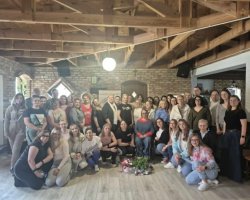 Zdjęcie grupowe kilkudziesięciu osób, głównie kobiet, stojących i kucających w przestronnej sali o rustykalnym wystroju. W tle widać ściany z jasnej cegły oraz odsłoniętą, drewnianą konstrukcję dachu. Uczestnicy są uśmiechnięci, ubrani w stroje codzienne. W centralnym punkcie, przed grupą, na podłodze leży bukiet kwiatów