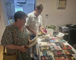 Kobieta w kwiecistej bluzce oraz mężczyzna w jasnej koszuli stoją przy białym stole w bibliotece, przeglądając i wybierając książki. Na blacie rozłożonych jest kilkadziesiąt różnych powieści oraz stojak z informacjami. W tle, na ścianie nad nimi, wisi klasyczny zegar wskazówkowy