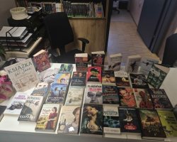 Widok z góry na biały stół w bibliotece, na którym równomiernie ułożono kilkadziesiąt książek popularnych autorów literatury obyczajowej, kryminalnej i sensacyjnej. Wśród nich wyróżnia się pionowo ustawiona tabliczka z napisem "Książka na św. Jana". W tle widoczne jest czarne biurowe krzesło obrotowe oraz regały z literaturą