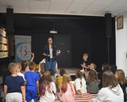 Kobieta w rozpiętej koszuli w kratę stoi na niskim podwyższeniu w sali bibliotecznej i trzyma retro projektor, przemawiając do grupy dzieci siedzących przed nią tyłem na pasiastym dywanie