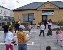 Grupa dzieci bawi się na wybrukowanym placu przed parterowym, żółtym budynkiem z szyldem „DOM HARCERZA”. Na nawierzchni placu widoczne jest namalowane białą farbą oznakowanie miejsca dla osób z niepełnosprawnościami